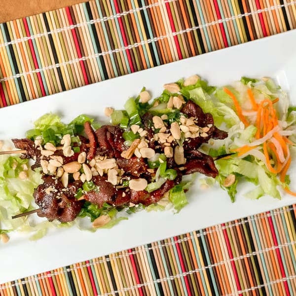 Two skewers of grilled meat topped with peanuts and green onions, served on lettuce with shredded carrots.
