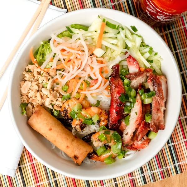 A Vietnamese vermicelli bowl with grilled pork, shrimp, egg roll, and vegetables on a colorful mat.