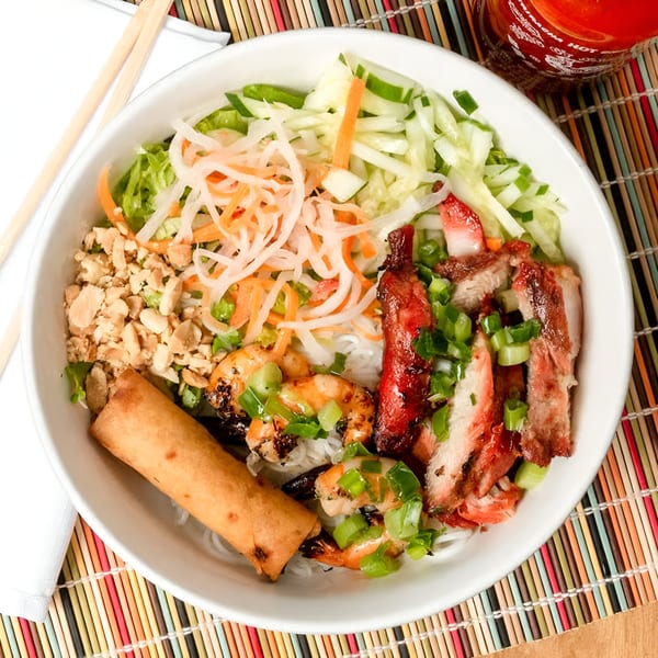 Vietnamese vermicelli bowl with grilled pork, shrimp, egg roll, and fresh vegetables, with Sriracha.