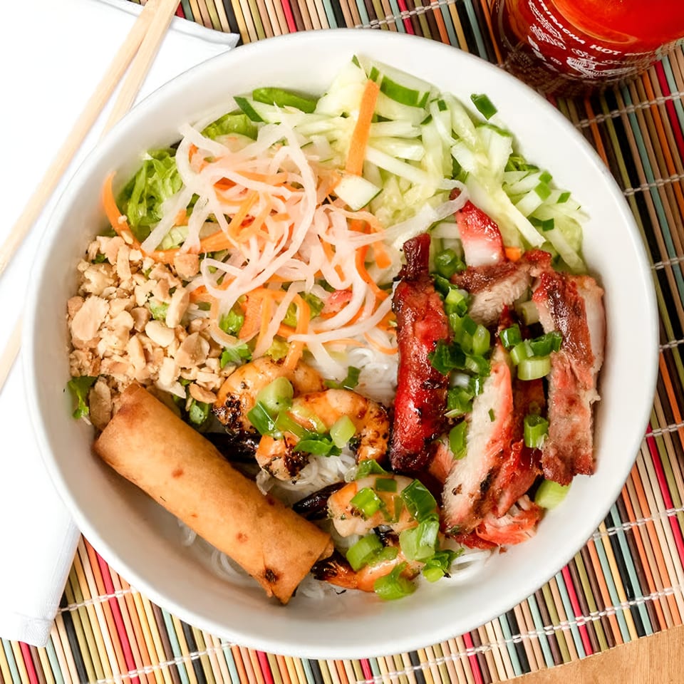 Vietnamese vermicelli bowl with grilled pork, shrimp, egg roll, and fresh vegetables, with Sriracha.